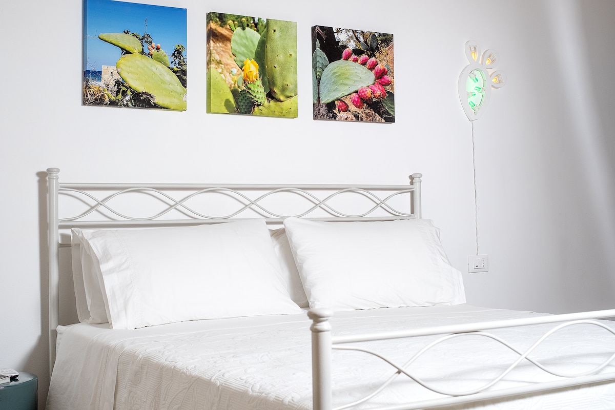 A white iron bed with fresh linens is positioned against a plain wall. Above the bed, three framed photographs of cacti are displayed, adding a touch of color. A softly glowing wall light is visible, contributing to the room's modern aesthetic.