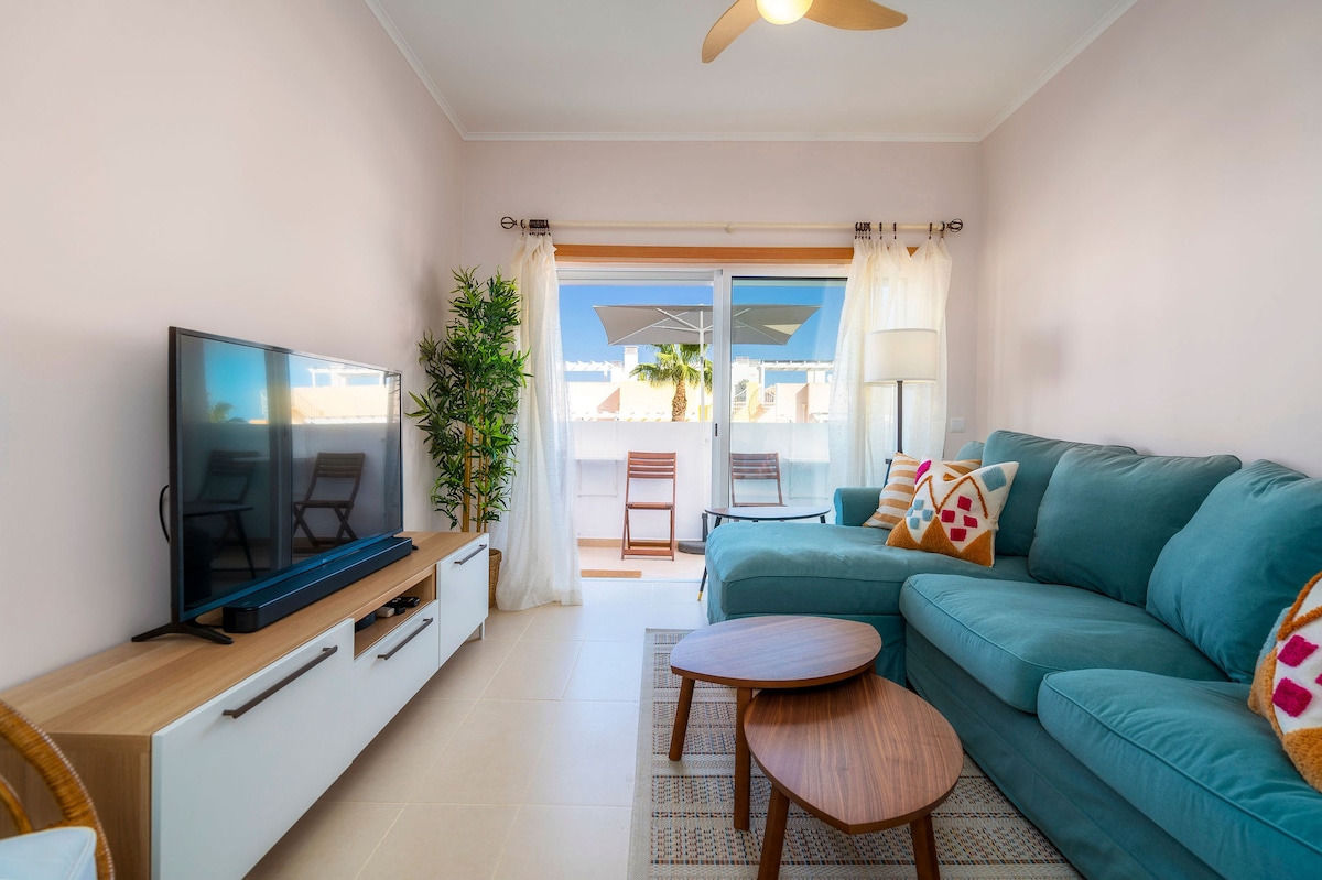 A comfortable living area features a plush sectional sofa in teal, complemented by wooden coffee tables. A flat-screen television is placed on a white entertainment unit. Sunlight filters through sheer curtains, illuminating the space that opens to a balcony with deck chairs.