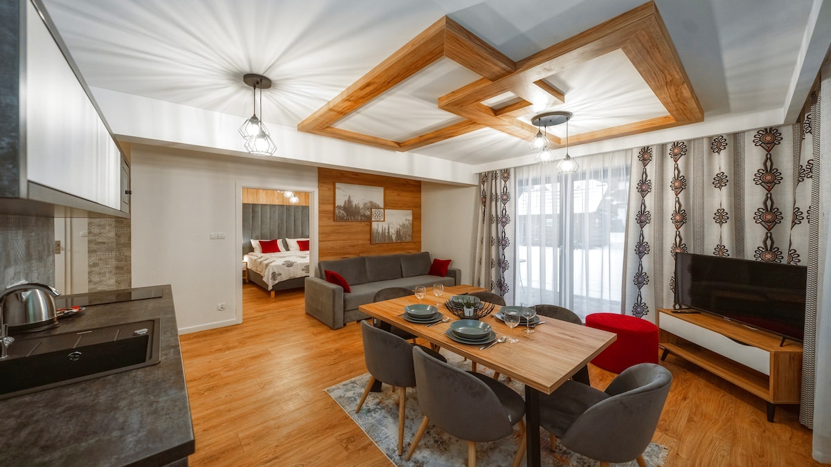 A spacious living area is highlighted by a wooden coffered ceiling. A dining table for six is centered among comfortable seating options. Large windows are adorned with patterned curtains, and a flat-screen TV is positioned near a cozy seating arrangement. A kitchen area is visible in the background.