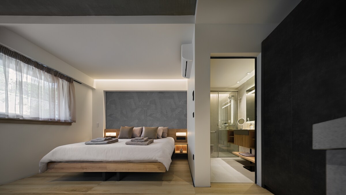 An open bedroom showcases a spacious double bed with neutral bedding and two decorative pillows. Soft lighting is present along the headboard, while a glass door leads to a private bathroom. Natural light filters in through sheer curtains, enhancing the minimalist design.