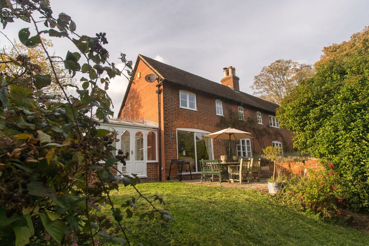 A charming two-story cottage presents a warm exterior with red brick and lush greenery. The patio area is equipped with a large umbrella providing shade over a dining table and chairs, offering a welcoming space for outdoor meals. The sunroom features expansive glass that allows natural light to fill the space.