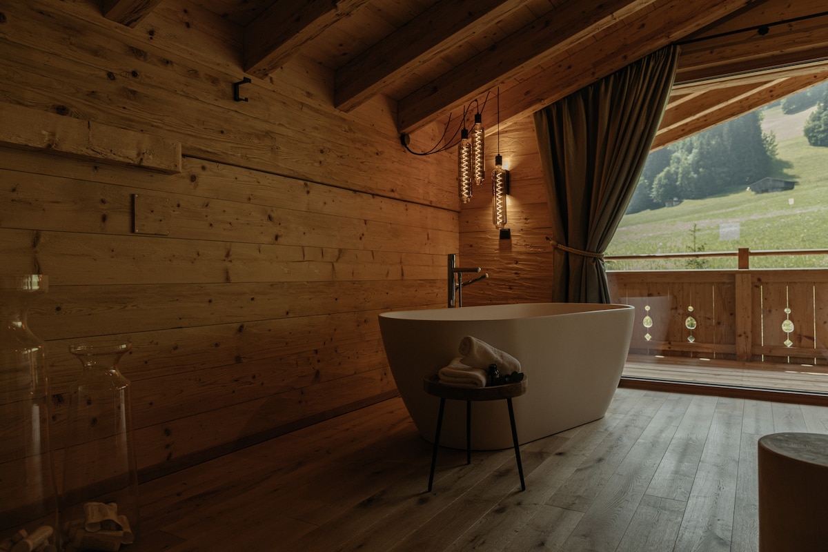 A modern freestanding bathtub is situated on wooden flooring beneath sloped ceilings. Natural light filters in through large windows, complemented by lush greenery outside. A small stool sits beside the tub, adding to the serene atmosphere of the wooden-paneled room.