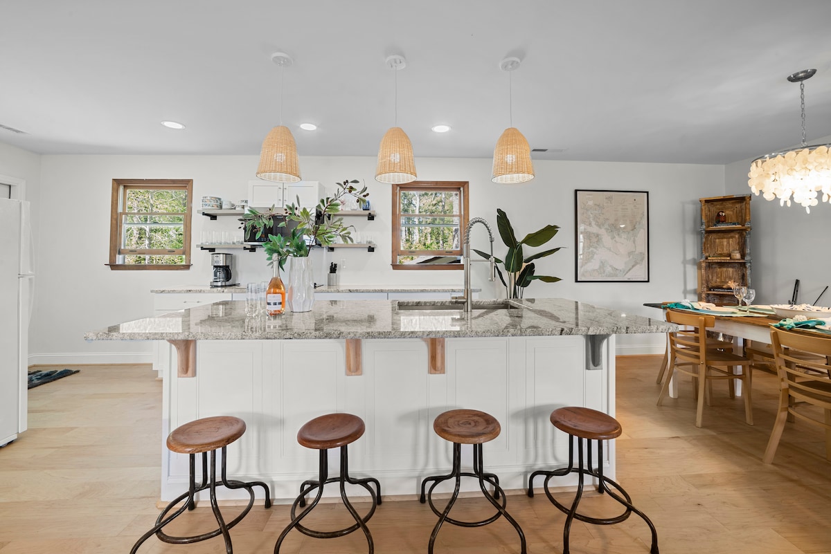 The kitchen features a large granite island with seating for four on wooden stools. Natural light enters through multiple windows, highlighting the minimalist design. A dining area is visible in the background, complemented by a cozy wooden table and lush greenery on display.