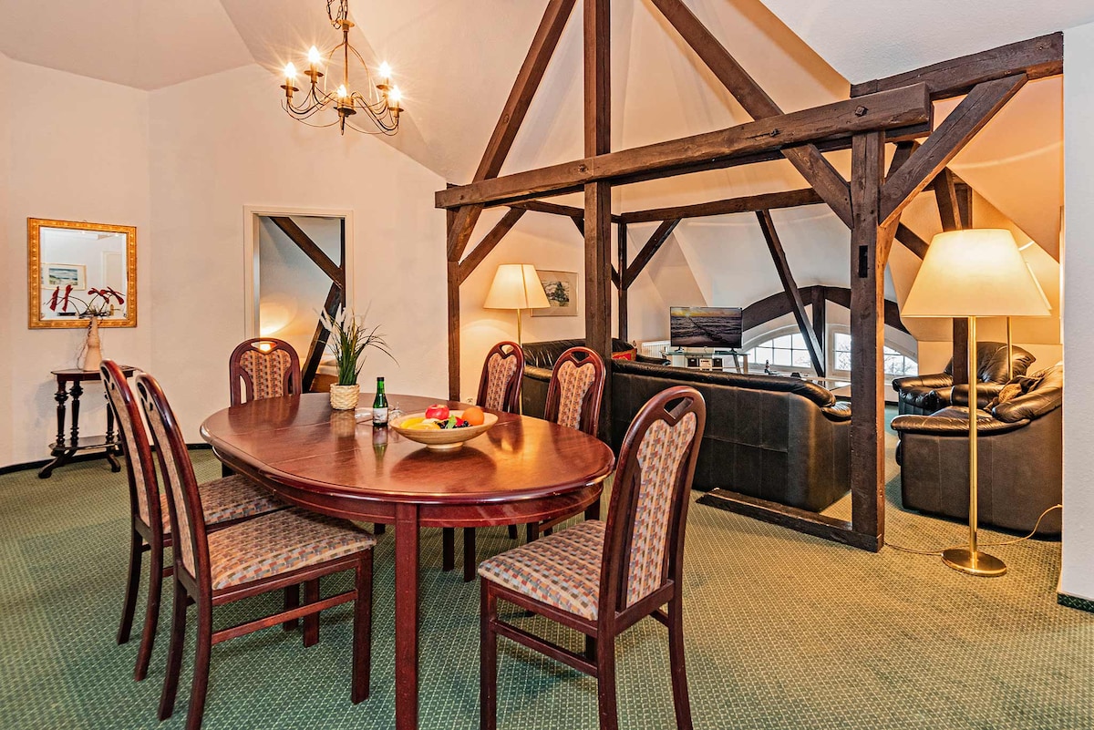 A spacious dining area is highlighted by a large wooden table surrounded by six patterned chairs. In the background, comfortable seating is arranged with a floor lamp and a TV visible, while wooden beams accentuate the high ceiling.