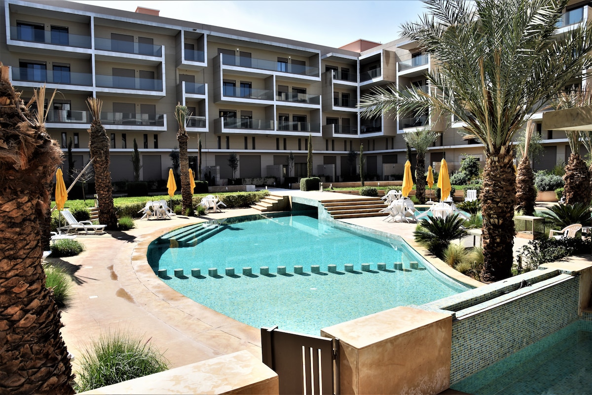 The image captures a tranquil pool area surrounded by palm trees and lounge chairs. Yellow umbrellas provide shade, while steps lead down to the inviting blue water. Multiple levels of greenery and landscaping enhance the serene environment of the community space.