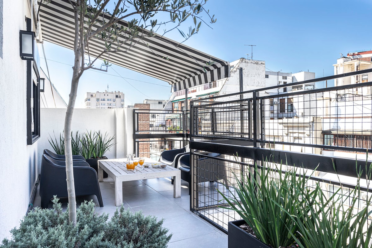 A private terrace is displayed, featuring modern furniture and a table set with drinks. Lush green plants line the edges, providing a touch of nature. A striped awning offers shade, while a clear blue sky completes the serene outdoor space.