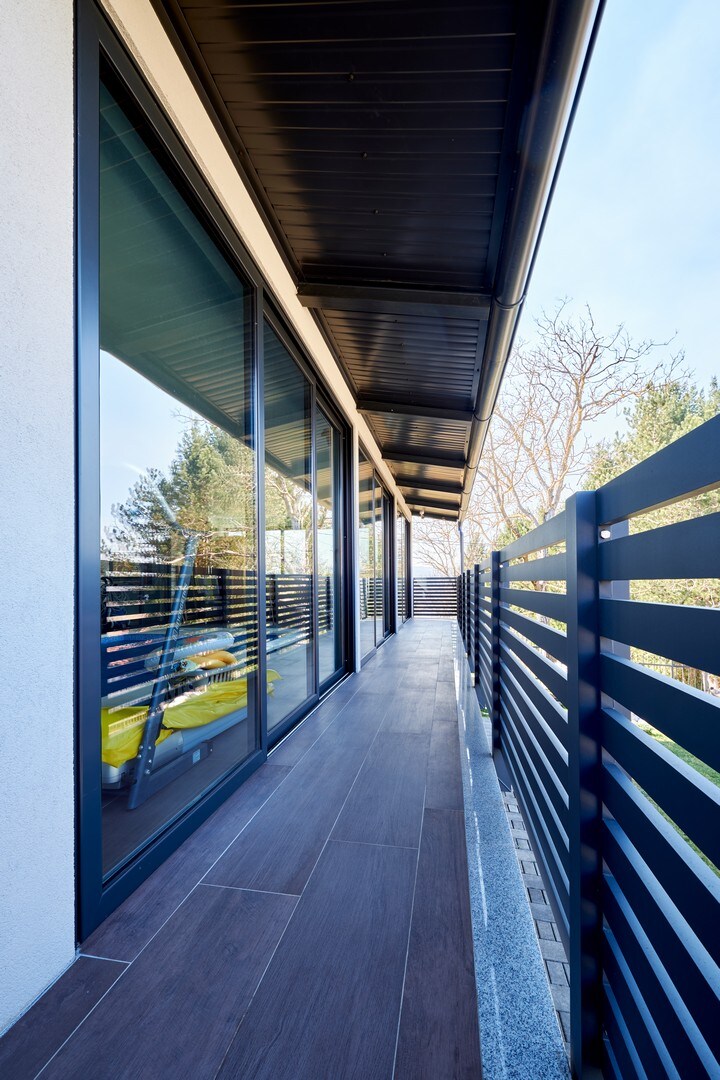 A long balcony is lined with black metal railing, providing a clear view of the surroundings. Large glass doors reflect the natural light and nearby greenery, while wooden flooring adds warmth to the outdoor space.