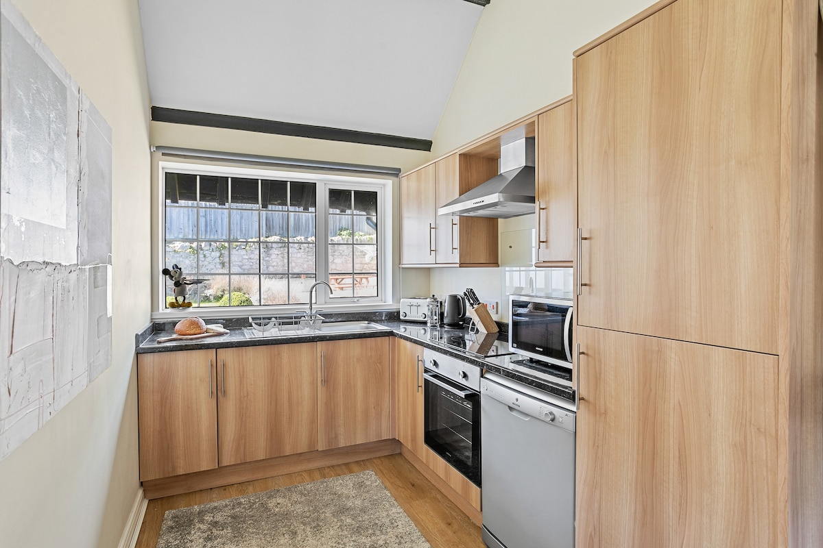 The kitchen offers a modern design with wooden cabinetry and granite countertops. Equipped appliances include a stove, oven, microwave, and refrigerator. Natural light pours in through a large window, illuminating the workspace and decorative elements throughout the area.
