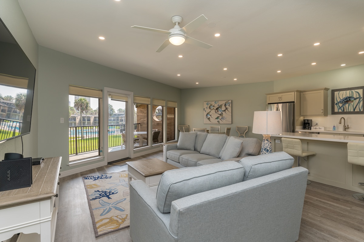The bright living area features a light grey sectional sofa and a wooden coffee table, positioned near large windows with views of the manicured grounds and pool. A flat-screen TV is mounted on the wall, and the open kitchen showcases modern amenities with neutral cabinetry.
