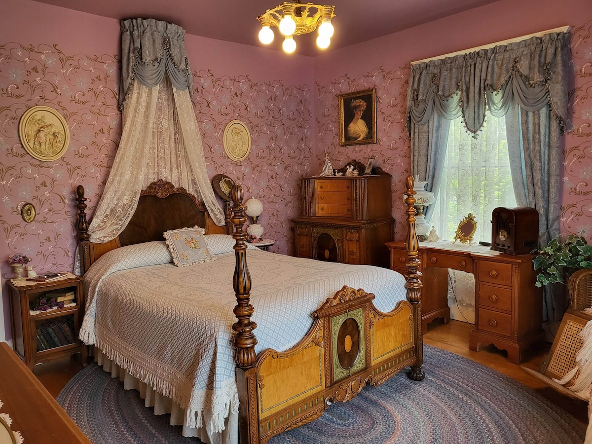 A charming bedroom features a four-poster bed with an ornate headboard, draped in delicate white linens. Vibrant pink wallpaper adorned with floral patterns creates an inviting atmosphere. A wooden dresser and desk complement the decor, illuminated by a vintage-style chandelier above.