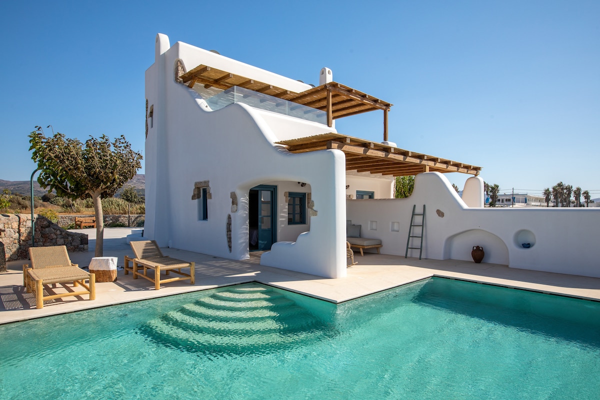 A whitewashed villa features a spacious outdoor area with a private pool. Sun loungers are positioned nearby, and the structure integrates natural elements with a wooden pergola. Lush greenery is visible in the background, enhancing the serene atmosphere of the property.