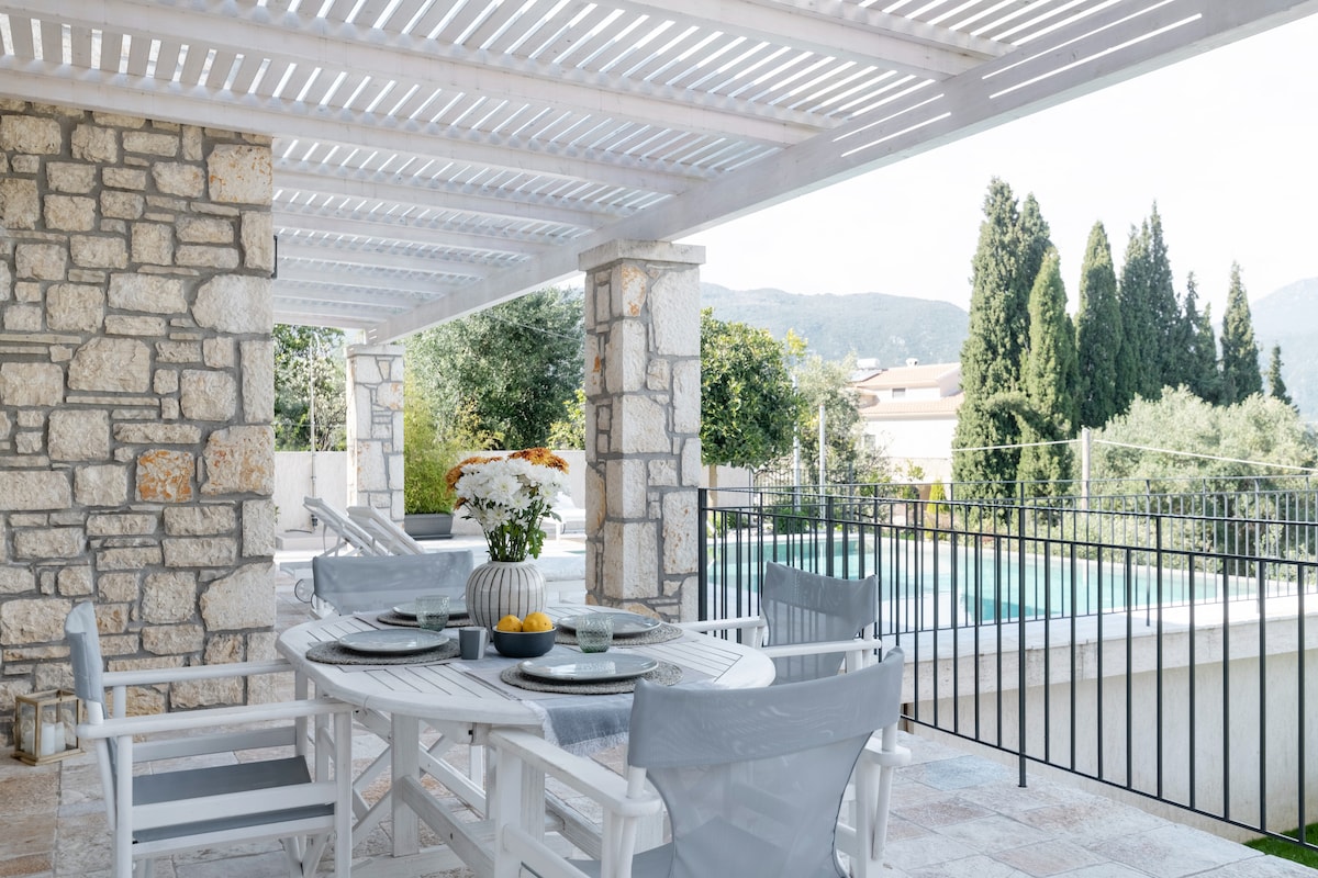 An outdoor dining area is presented under a slatted pergola, featuring a large table set for meals. Comfortable seating is arranged nearby, with a view of a private pool and lush greenery in the background. The mountains are faintly visible beyond the trees.