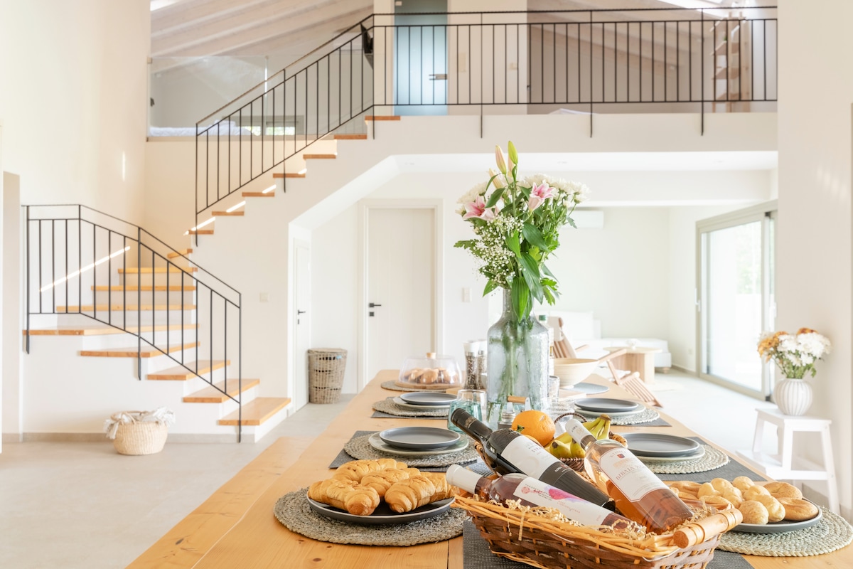 A dining area features a long wooden table set with plates, glassware, and a basket of baked goods. A floral arrangement adds a touch of color. Stairs lead to an upper level, while glass doors open to an outdoor space, highlighting the villa's airy design.