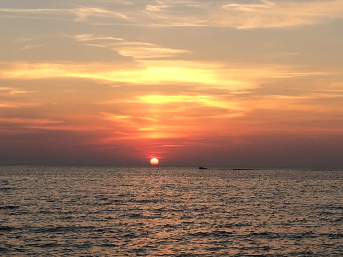 A serene sunset casts warm hues of orange and pink over the calm waters, with the sun dipping below the horizon. A small boat is visible in the distance, contributing to the tranquil beach atmosphere.