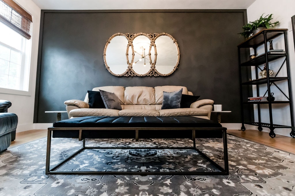 A warmly decorated living area features a light-colored sofa with decorative pillows, centered on a patterned area rug. Two ornate mirrors are mounted on a dark accent wall, reflecting light. A shelving unit stands to one side, holding various decorative items.