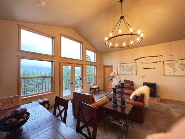 A spacious living area is highlighted by large windows offering views of the surrounding landscape. A circular chandelier hangs above a wooden dining table and dark leather seating. Decorative maps and vintage rifles are displayed on the walls, adding character to the room.