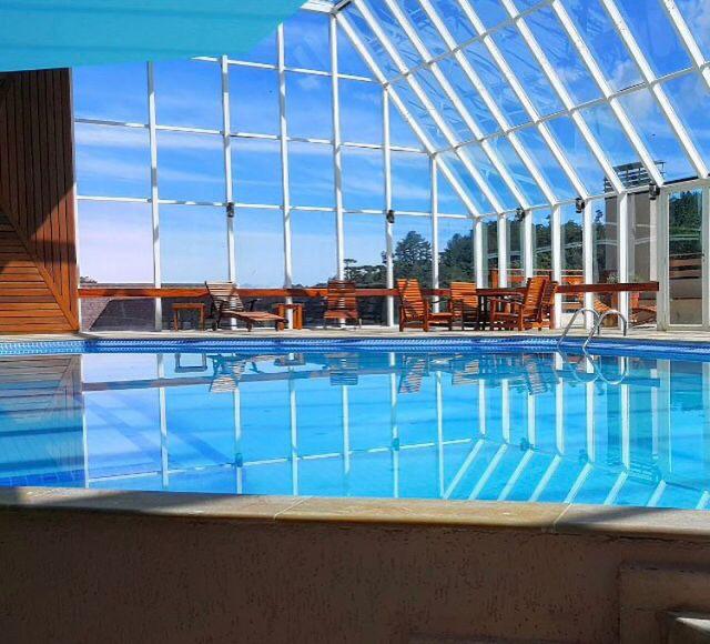An indoor swimming pool is showcased under a glass roof, allowing natural light to illuminate the clear water. Surrounding the pool are wooden lounge chairs, creating a comfortable area for relaxation. The scenic view through the glass panels enhances the tranquil atmosphere.