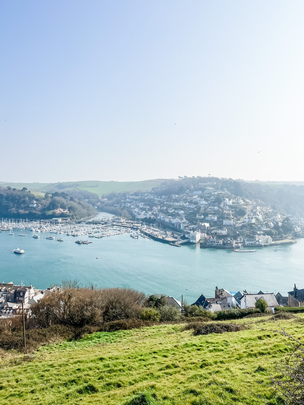 The image presents a scenic view of a tranquil waterway, dotted with boats. Surrounding hills display vibrant greenery, complemented by quaint buildings lining the riverbank. The clear blue sky enhances the serene atmosphere, creating a picturesque backdrop of the coastal landscape.