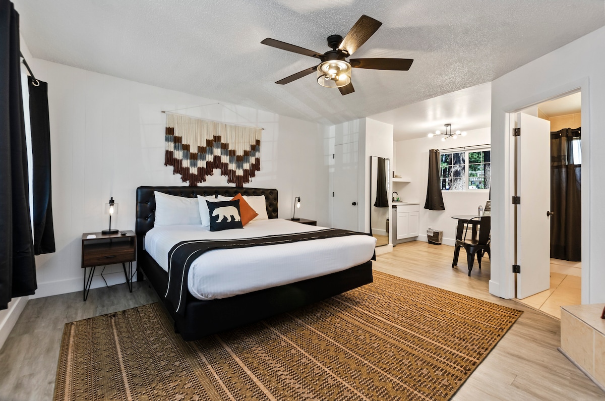 A spacious room features a king bed with a patterned throw pillow, complemented by a cozy area rug. A ceiling fan and bedside lamps provide lighting. The adjoining bathroom includes a vanity and a shower, while large windows allow natural light to enhance the space.