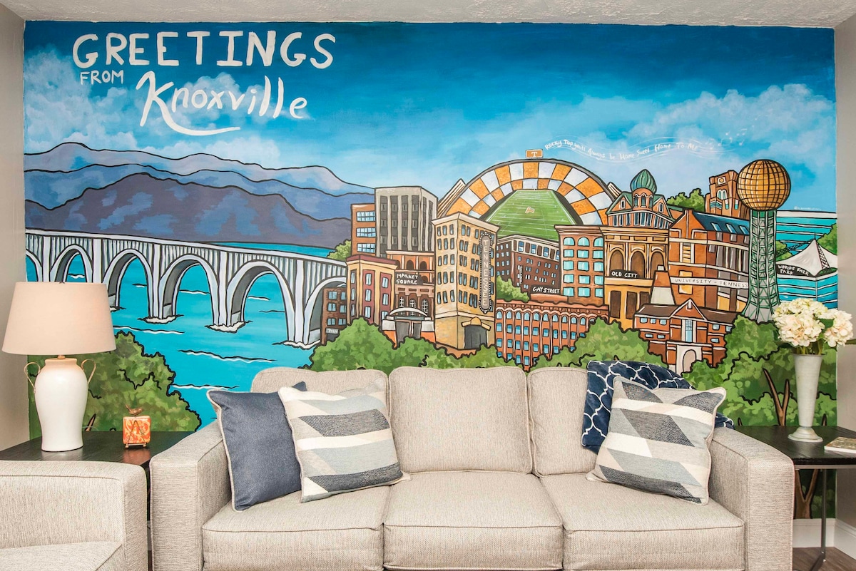 A living area features a large mural depicting a vibrant cityscape of Knoxville, complete with the iconic Sunsphere and the Tennessee River. A light-gray sofa adorned with striped and solid blue pillows is positioned nearby, complemented by a lamp casting a warm glow.