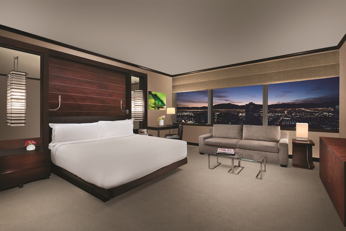 A spacious studio suite features a king bed with crisp white linens. A comfortable sofa and modern side tables are arranged near floor-to-ceiling windows, offering stunning views of the city skyline. Soft lighting enhances the welcoming ambiance.