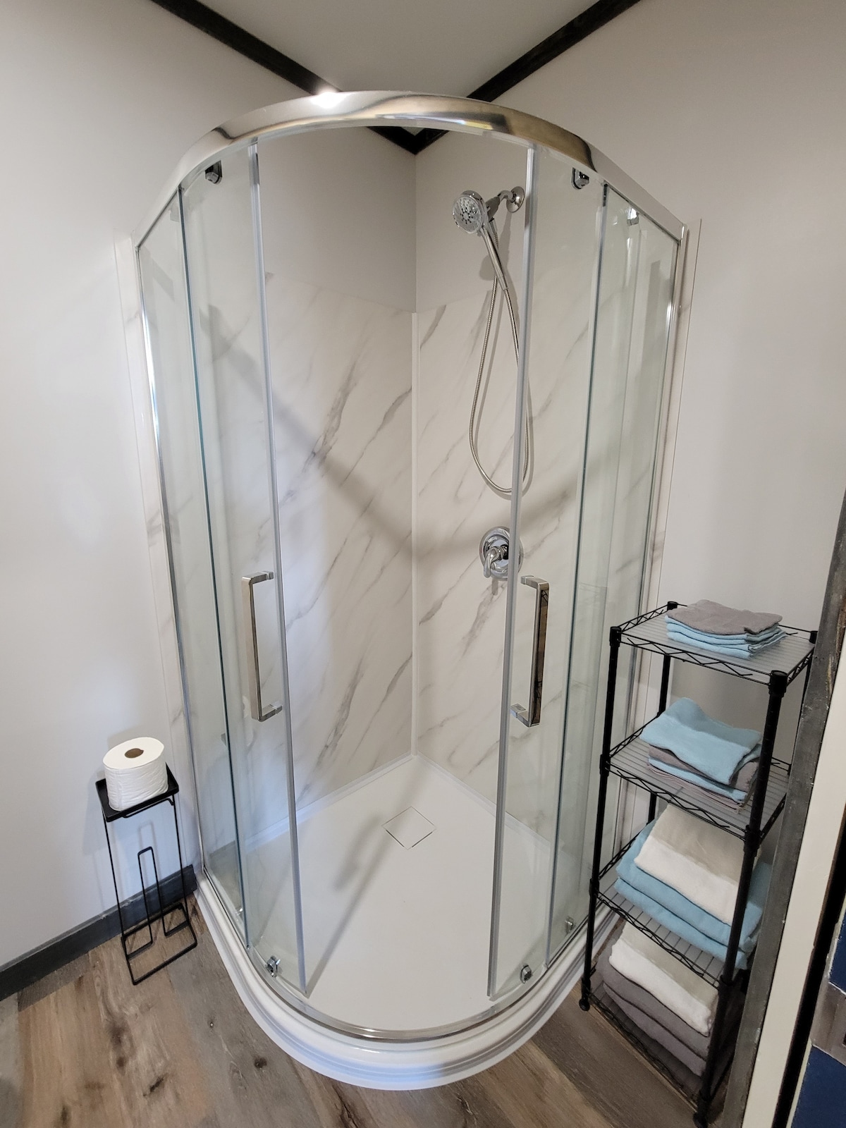 A modern shower stall is enclosed with clear glass panels, showcasing a sleek design featuring a rainfall showerhead and a built-in corner shelf. Soft light reflects off the white marble-patterned walls, while a small stand holds neatly stacked towels in various shades, alongside a toilet paper roll.