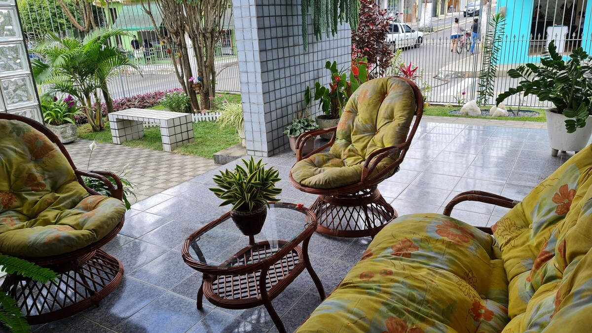 A covered outdoor seating area is furnished with two cushioned rocking chairs and a coffee table. Lush greenery and colorful flowers surround the space, creating a refreshing atmosphere. The tiled floor leads to a view of the garden and pathway beyond.