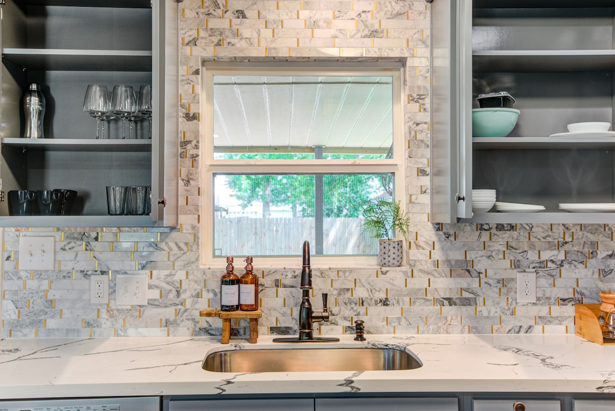 The kitchen features a sleek counter with a modern sink, complemented by a mosaic tile backsplash. Glassware is neatly arranged on open shelves, while natural light filters through a window, offering a view of the backyard. Simple decor elements are present, enhancing the functionality of the space.