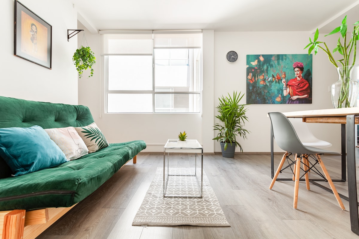 A vibrant living area is highlighted by a green sofa bed, accompanied by decorative cushions. Natural light enters through large windows, illuminating a small coffee table and a patterned area rug. A potted plant and wall art add a touch of warmth to the space.