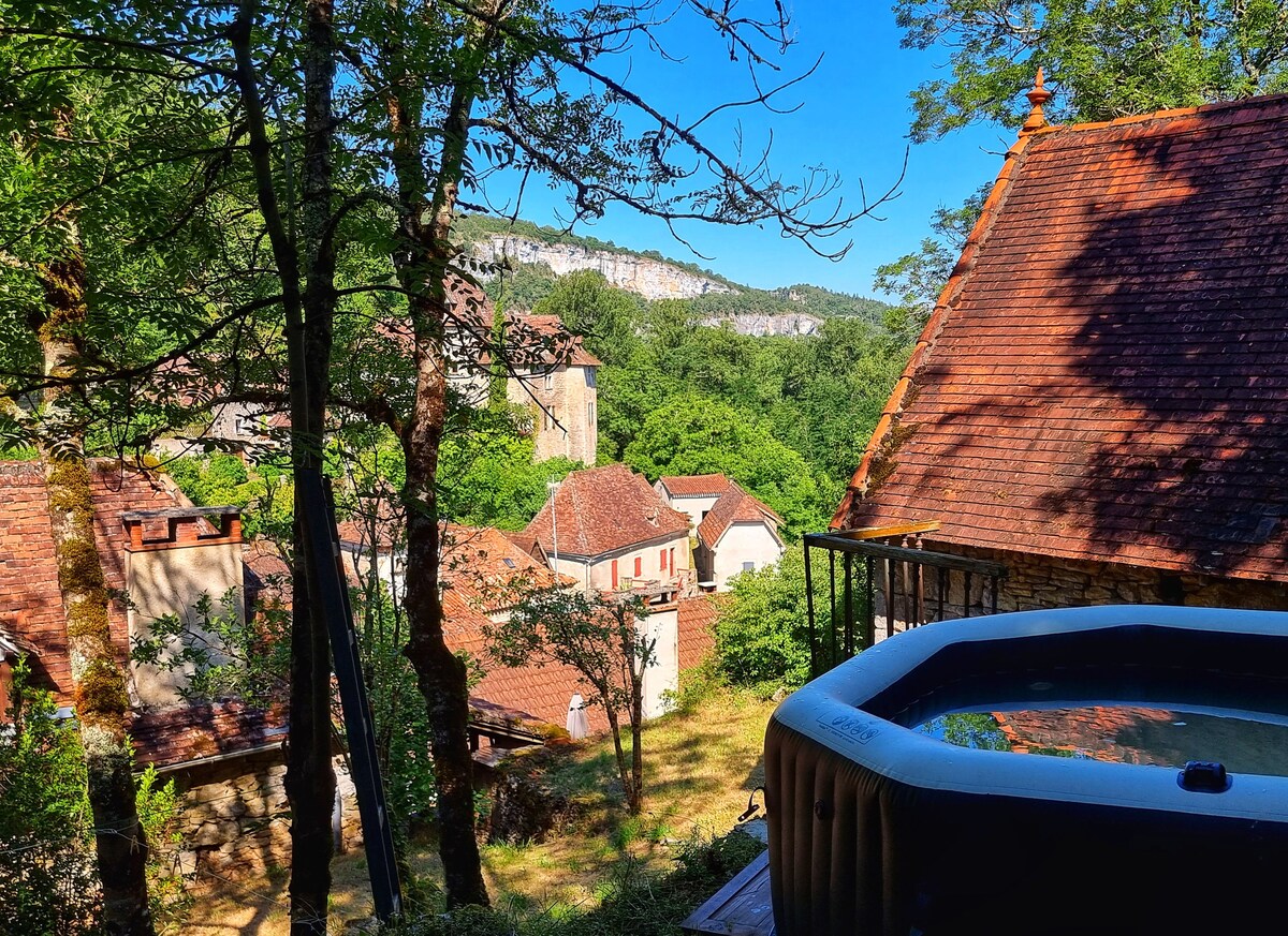 An outdoor terrace view reveals a relaxing hot tub surrounded by trees. Below, charming village rooftops can be seen, framed by lush greenery and distant rugged hills under a clear blue sky.