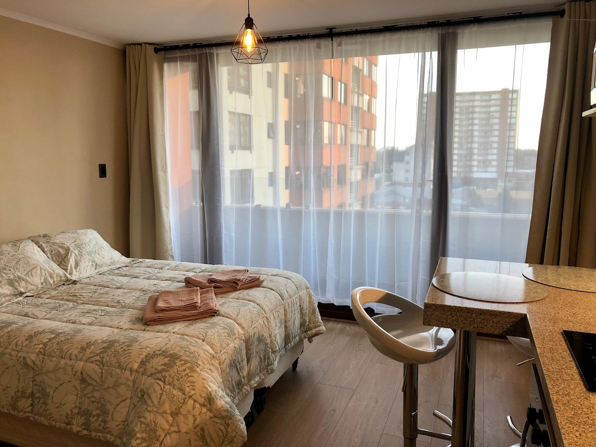 A spacious sleeping area is furnished with a bed featuring a floral-patterned coverlet. Large floor-to-ceiling windows are adorned with sheer curtains, allowing natural light to fill the room while providing a view of the surrounding cityscape.