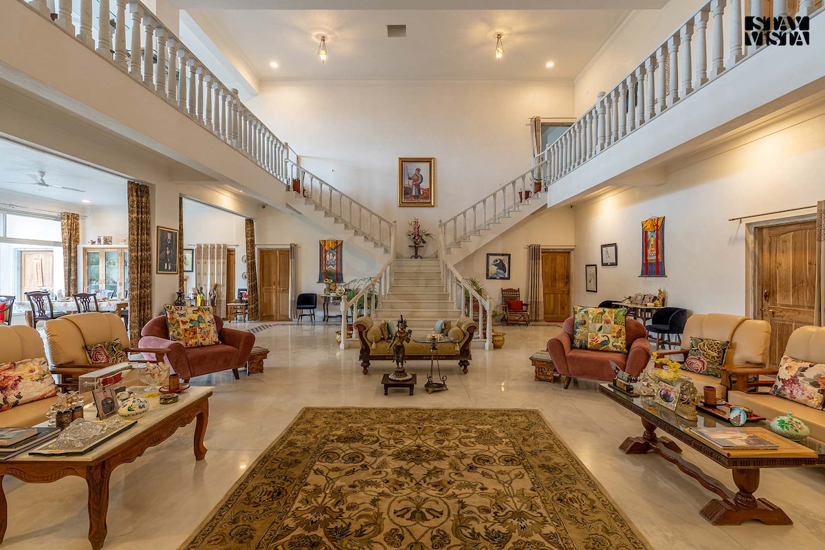 An expansive living area features high ceilings and a grand staircase at its center. Comfortable seating arrangements with floral patterns are arranged alongside elegant wooden furniture. Decorative rugs and art pieces add character to the space, while large windows allow natural light to fill the room.