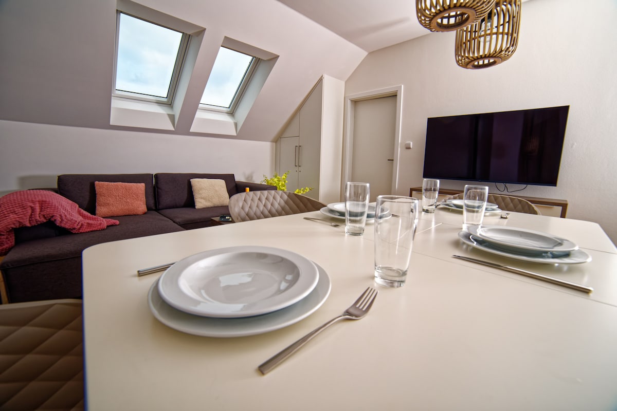 A modern dining area features a contemporary table set for four, including plates and glasses. A comfortable sofa with colorful cushions is visible in the background, alongside sloped skylights that allow natural light to fill the space. A wall-mounted TV adds convenience to the room.