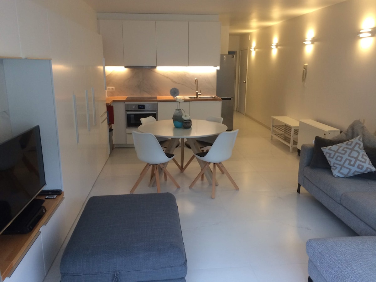 A modern living area is displayed, featuring a round dining table surrounded by white chairs. A sleek kitchen with marble countertops is visible in the background. A gray sofa and matching ottoman provide comfortable seating, complemented by soft lighting along the walls.