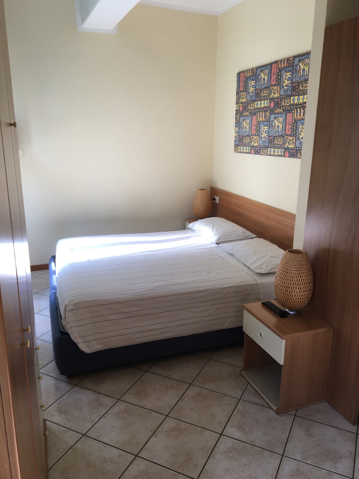 A compact sleeping area is furnished with a spacious bed, topped with white linens. Flanking the bed are two decorative lamps placed on side tables. A colorful wall art piece adds character to the light-colored walls, while natural light enters through a nearby window.