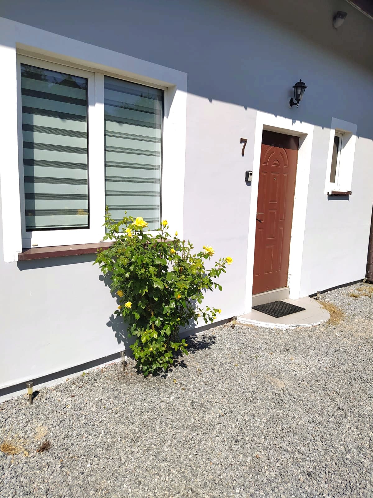 An entrance to the home is presented, featuring a brown door set within a light-colored wall. A potted plant with yellow flowers adds a touch of color beside the door, while a gravel pathway leads to the entrance, framed by a window with horizontal blinds.