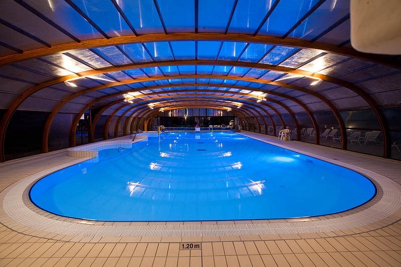 An indoor swimming pool is featured, enclosed by a wooden structure with a transparent roof. The water appears invitingly blue, and the space is softly illuminated, creating a calm ambiance. Lounge chairs are arranged around the poolside, enhancing the relaxation area.