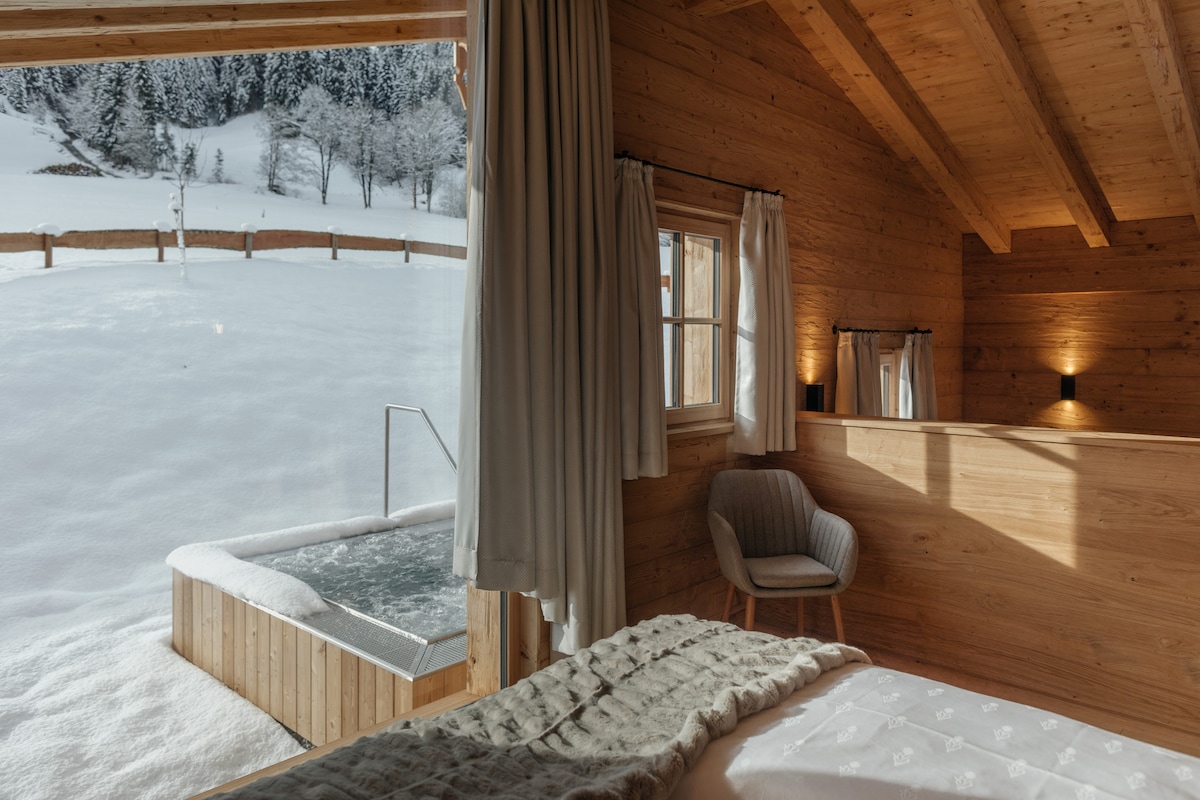 A serene bedroom features wooden walls and ceilings, illuminated by soft lighting. A large window provides a view of the snow-covered landscape outside, alongside a private wooden hot tub. A cozy chair is positioned in the corner, with light fabrics adorning the spacious bed.