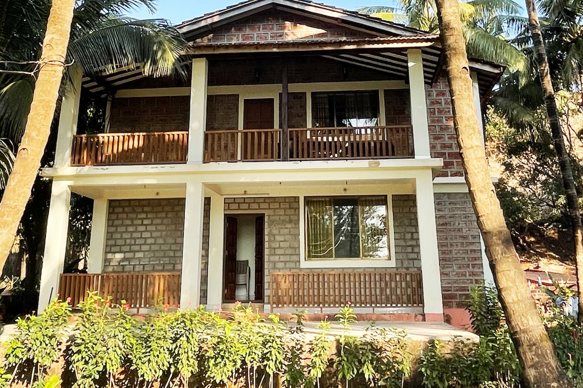 A two-story house features a stone façade with wooden balcony railings. Lush greenery surrounds the building, and tall palm trees are visible nearby. Each floor has a covered porch area, while large windows allow natural light into the rooms.