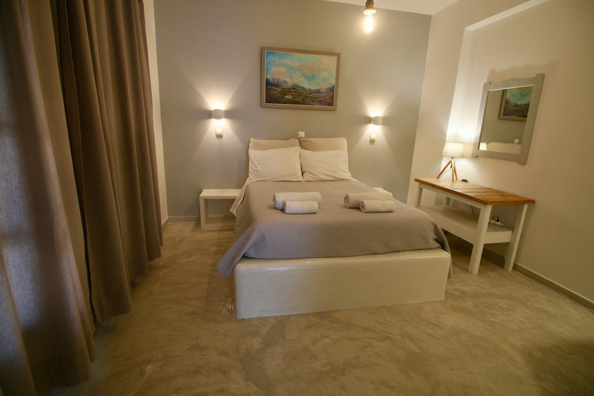 A guest room features a comfortable bed dressed in neutral linens, complemented by two decorative towels. Soft lighting is provided by wall sconces on either side. A wooden nightstand and a mirror enhance the space, while large curtains allow for natural light.