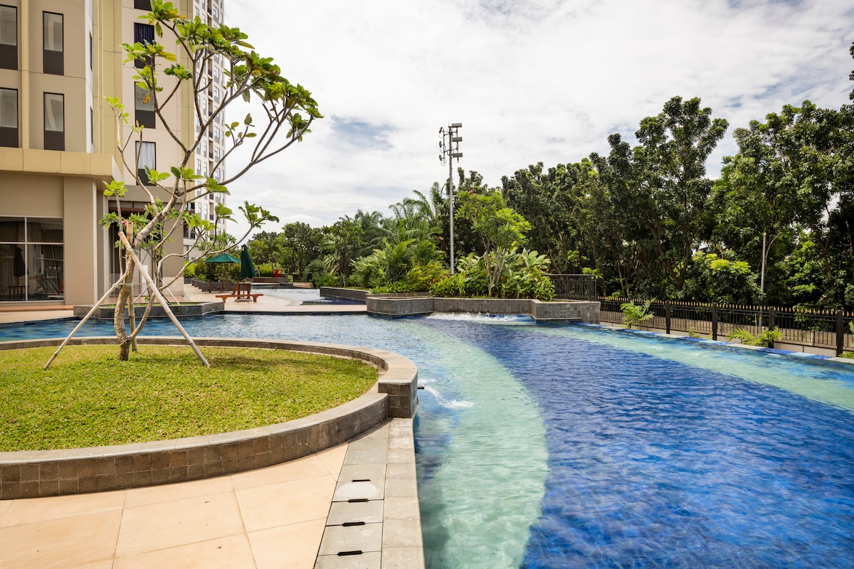 A serene outdoor area features a winding swimming pool surrounded by lush greenery. A manicured lawn with a small tree adds a natural touch, while the contemporary building stands in the background. The scene is brightened by soft, natural sunlight.