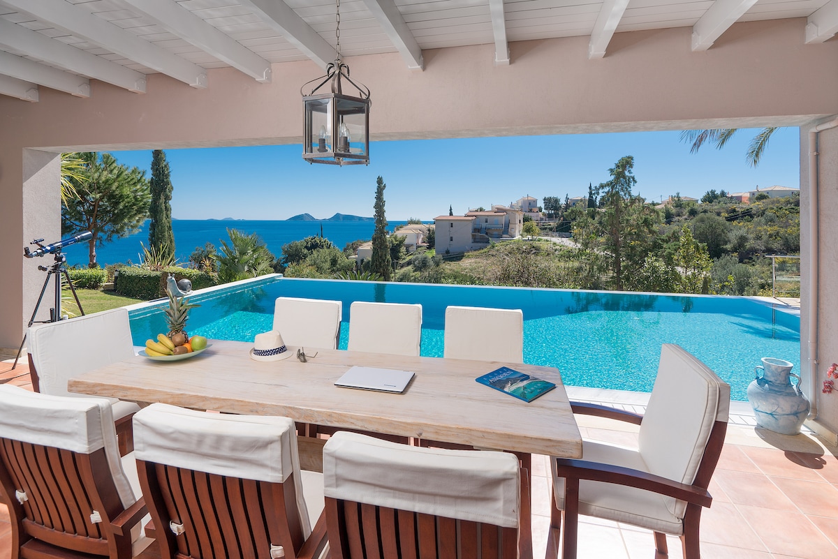 An outdoor dining area features a long wooden table surrounded by comfortable chairs, positioned under a covered terrace. Beyond, an infinity pool extends towards the sea, reflecting the clear blue sky. A picturesque view of the landscape and distant islands is visible in the background.