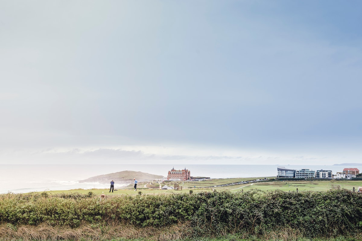 A distant view showcases the coastline, with rolling green fields leading to the ocean. The iconic Headland Hotel is prominently featured, surrounded by serene landscapes. The sky transitions from soft blues to grays, hinting at the coastal ambiance of Fistral Beach.