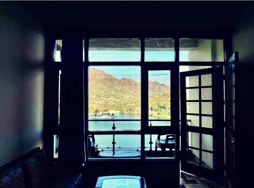 A large window frames a view of the serene lake and rolling hills beyond. Natural light fills the room, highlighting the seating area with a couch and coffee table. The door is open, inviting the outdoor scenery into the space.