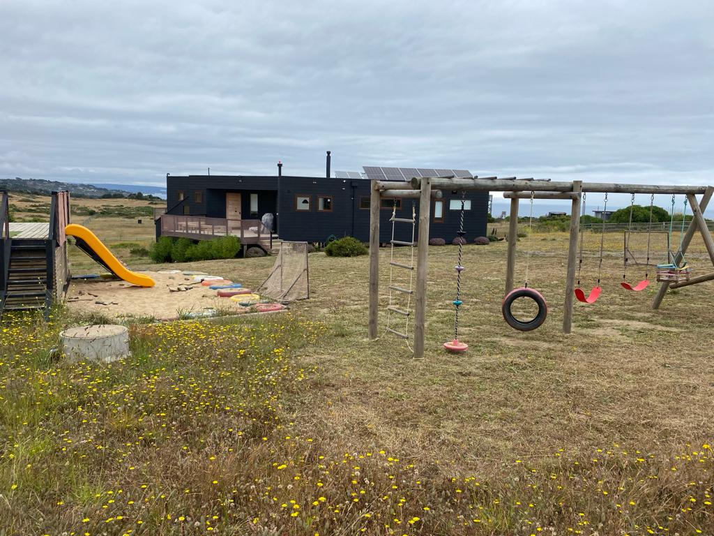 A playground area is visible with a slide, swings, and climbing structures, set against a backdrop of a black house featuring solar panels. Natural grass and wildflowers cover the ground, creating a serene outdoor space. The surrounding landscape showcases open fields and a cloudy sky.