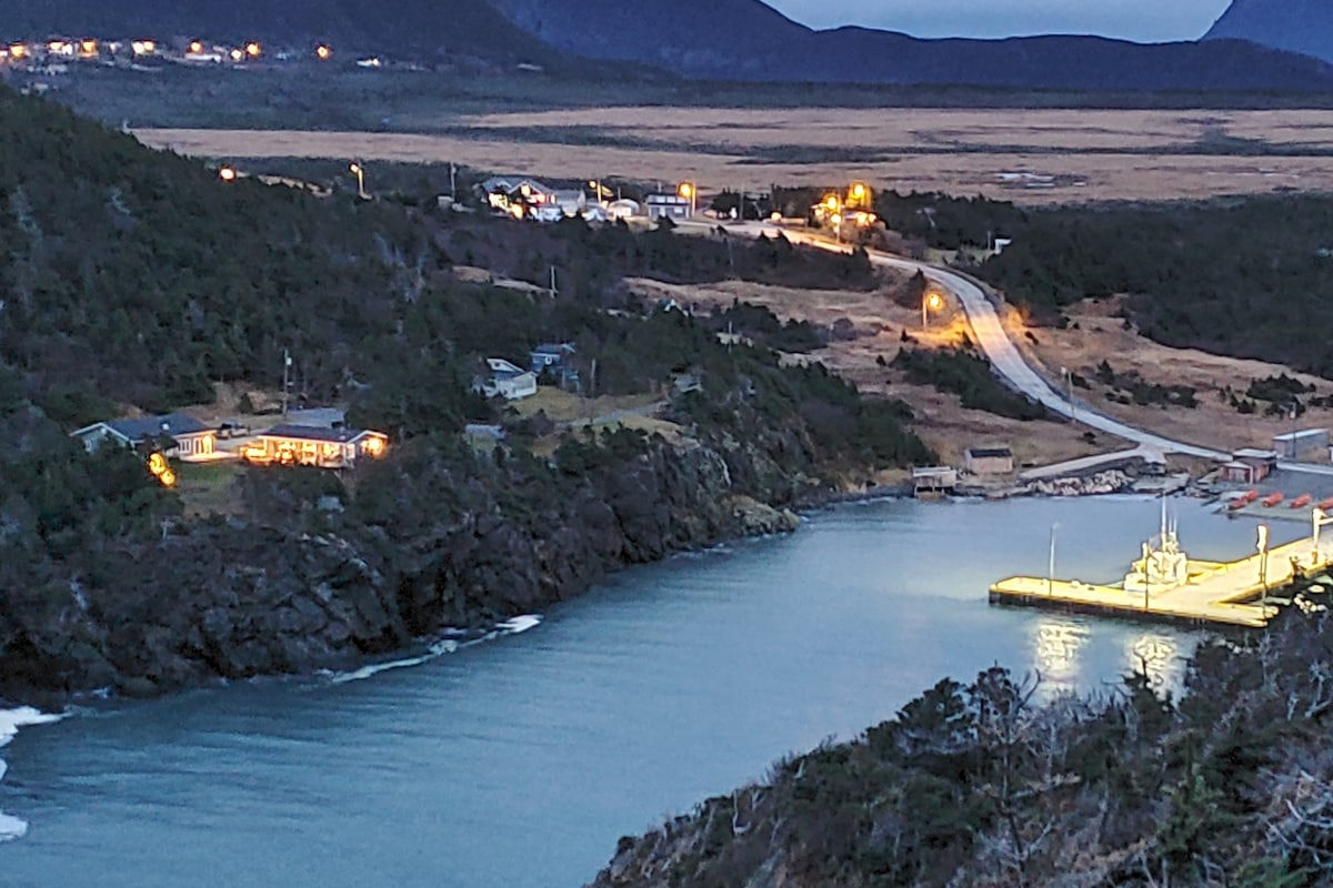 A serene evening view of Little Port, highlighted by gently glowing lights from nearby buildings. The calm water reflects the lights, and the rugged landscape transitions from the illuminated dock to the surrounding cliffs leading up to distant mountains.