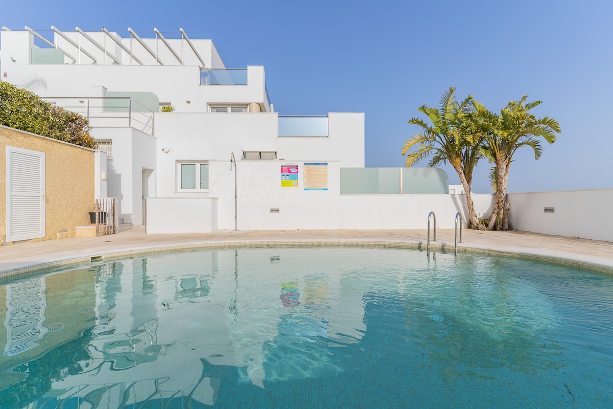 A communal pool is featured, surrounded by a smooth deck and palm trees. The clear water reflects the blue sky, with a stair entry visible. White buildings are positioned in the background, showcasing contemporary architectural design.