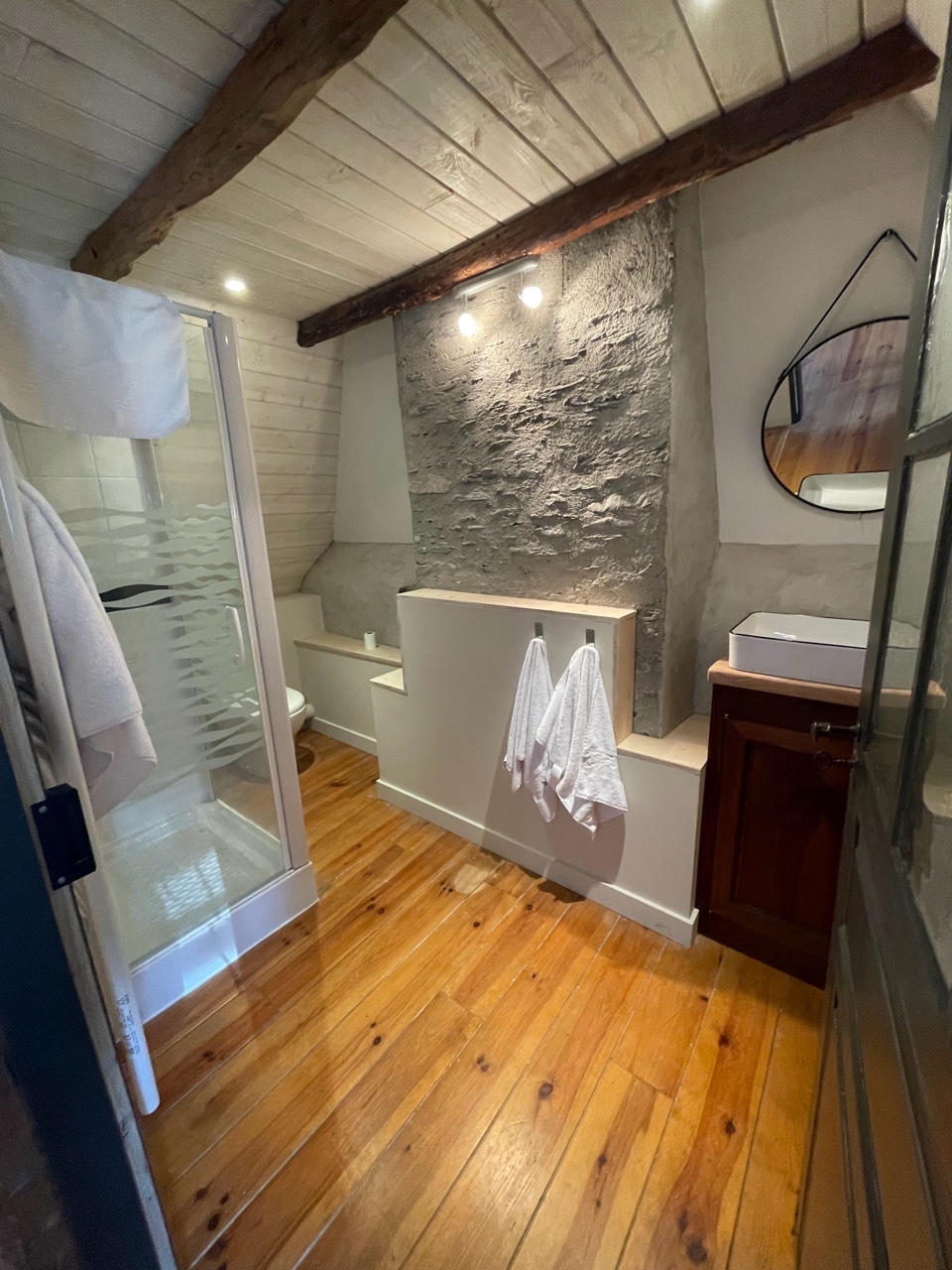 The bathroom features rustic wooden flooring and exposed beams, providing a cozy feel. A modern shower stall is situated beside a wooden vanity with a mirror above. Two towels are neatly hung on a wall-mounted rack, enhancing the functional design of the space.