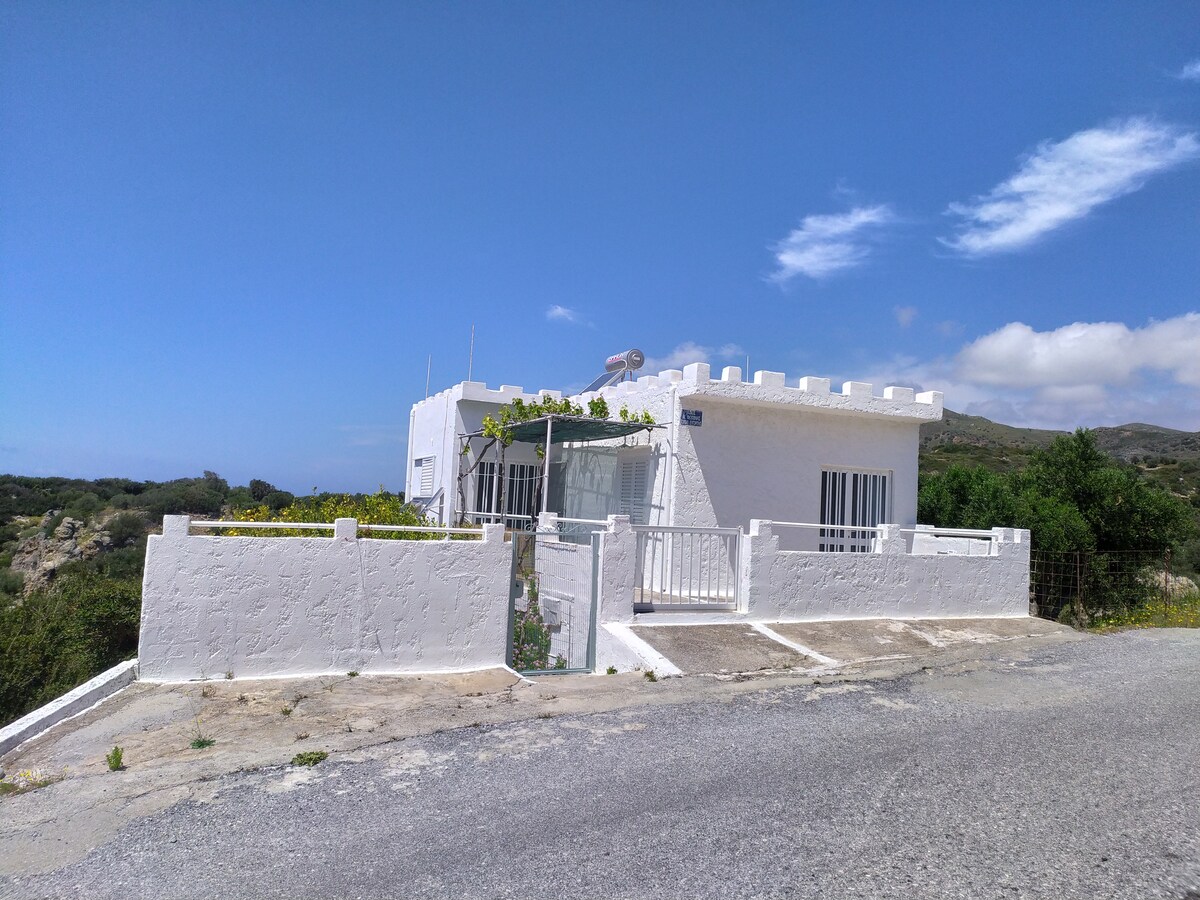 A white house with a glass entrance is situated on a hillside, surrounded by greenery and open fields. The structure features large windows and a gated front yard, with a scenic view of the landscape and sky beyond.