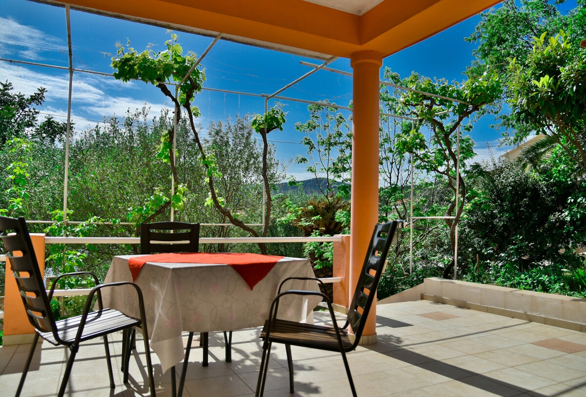 A covered outdoor patio is presented, featuring a table set with a vibrant red tablecloth and surrounded by four sturdy chairs. Green vines are seen climbing along the structure, and lush greenery filled with various plants is visible in the background, enhancing the natural environment.